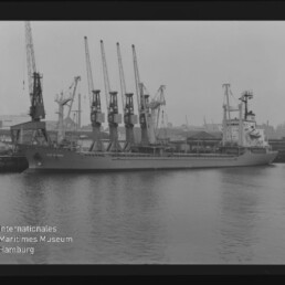 Schwarzweißfoto eines am Kai liegenden Frachtschiffs mit vier Ladekränen und dem Schriftzug Maritimes Museum Hamburg.