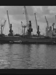 Frachtschiff am Kai vor einer Reihe Hafenkräne, ruhiges Wasser im Vordergrund und bewölkter Himmel, Museumslogo unten links.