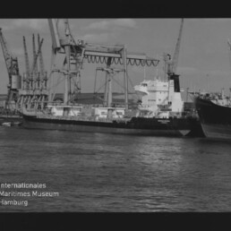 Frachtschiff am Kai vor großen Hafenkränen, Wasser im Vordergrund; Schriftzug Internationales Maritimes Museum Hamburg