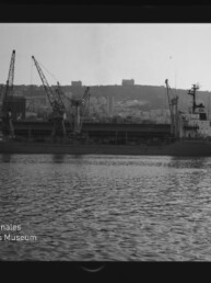 Frachtschiff am Kai neben mehreren Hafenkränen, dahinter Stadt auf einem Hügel und spiegelndes Wasser im Vordergrund.
