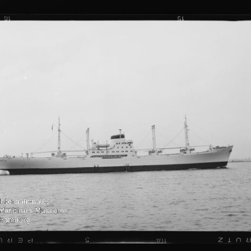 Seitliche s/w-Ansicht eines großen Frachtschiffs mit Aufbauten und Ladekränen, ruhig auf dem Wasser; Museumslogo unten links.