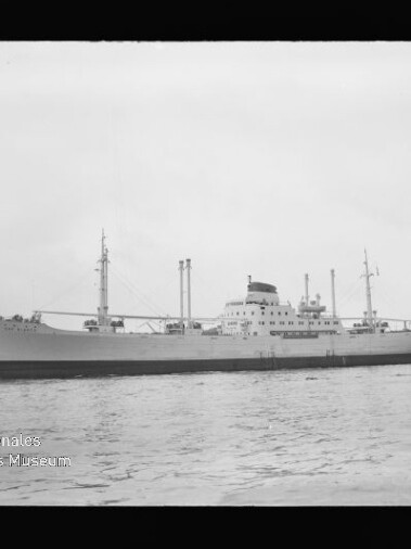 Schwarzweiß-Foto eines großen Frachtschiffs in Seitenansicht mit Mittschiffsaufbau, mehreren Masten und Schornstein, ruhig im Hafen liegend