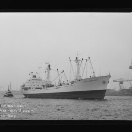 Großes Frachtschiff mit Ladebäumen, von einem Schlepper begleitet, vor Industriehafen mit hohen Portalkränen.