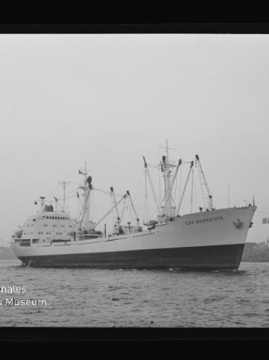 Großes Frachtschiff mit Ladebäumen, von einem Schlepper begleitet, vor Industriehafen mit hohen Portalkränen.