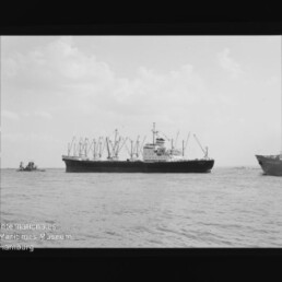 Frachtschiff mit mehreren Ladekränen auf ruhiger See, Kleinfahrzeuge am Rand; Stempel Maritimes Museum Hamburg.
