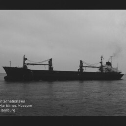 Schwarzweiß-Silhouette eines Frachtschiffs mit zwei Ladekränen auf ruhiger See; Museumslogo Internationales Maritimes Museum Hamburg sichtbar.