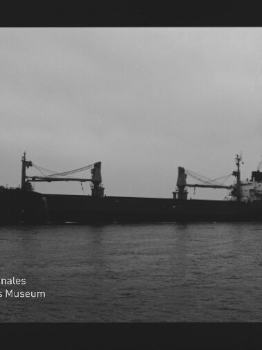 Schwarzweiß-Silhouette eines Frachtschiffs mit zwei Ladekränen auf ruhiger See; Museumslogo Internationales Maritimes Museum Hamburg sichtbar.
