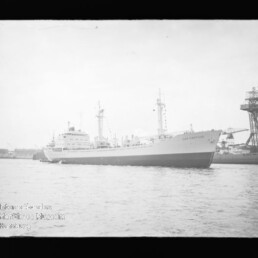 Schwarzweißaufnahme eines großen Frachtschiffs in Seitenansicht im Hafen, rechts ein hoher Werftkran und Industrieanlagen.