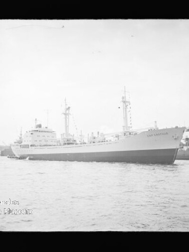 Schwarzweißaufnahme eines großen Frachtschiffs in Seitenansicht im Hafen, rechts ein hoher Werftkran und Industrieanlagen.