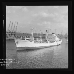 Weißes Frachtschiff mit mehreren Masten und Ladekränen im Hintergrund liegt im Hafen bei ruhigem Wasser und bewölktem Himmel.