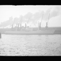 Dampfiges Schwarz-Weiß-Foto eines Frachtschiffs am Industriehafen mit Kränen im Hintergrund und einer Boje im Vordergrund.