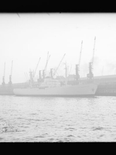 Weißes Schiff am Industriehafen mit einer Reihe hoher Kräne im Nebel, Wasser im Vordergrund und Museumsstempel links unten.