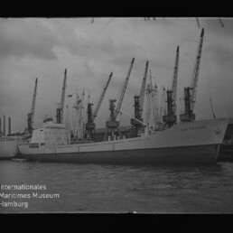 Frachtschiff am Kai, mehrere Hafenkräne heben sich im Hintergrund, ruhige Wasserfläche im Vordergrund.