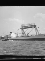 Großes Frachtschiff fährt vor einer Werft mit hohem Portalkran und mehreren Kränen; kleines Motorboot im Vordergrund.