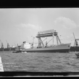 Großes Frachtschiff fährt vor einer Werft mit hohem Portalkran und mehreren Kränen; kleines Motorboot im Vordergrund.