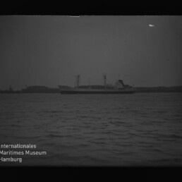 Entferntes Frachtschiff mit Ladekränen vor flachem Küstenhorizont, ruhige See und Museumswasserzeichen unten links.