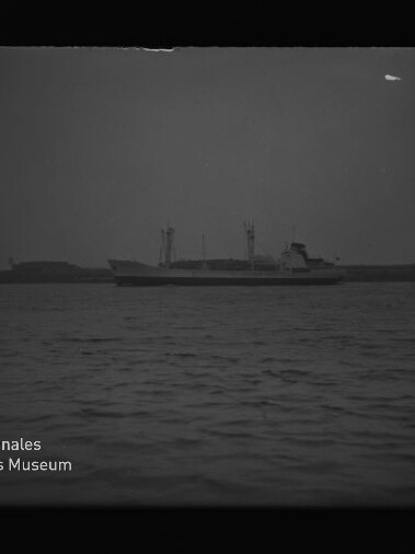 Entferntes Frachtschiff mit Ladekränen vor flachem Küstenhorizont, ruhige See und Museumswasserzeichen unten links.