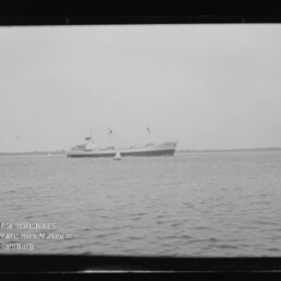 Schiff mit zwei Masten auf ruhigem Meer vor flachem Horizont, Boje im Vordergrund und Museumssiegel unten links.