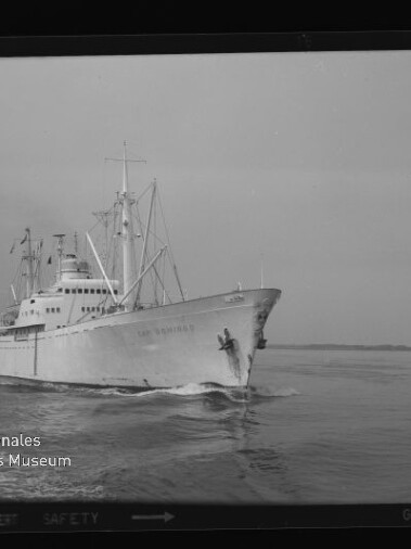 Weißes Frachtschiff mit Aufbauten und Ladekränen fährt bei ruhiger See, von vorn links aufgenommen.