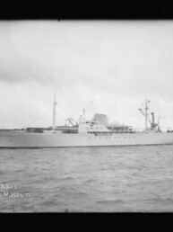 Weißes Passagierschiff in Seitenansicht auf dem Wasser vor Hafenkränen, Museumslogo links unten.
