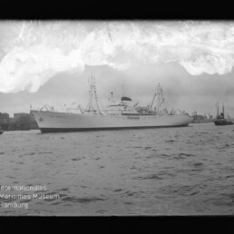 Weißes Passagierschiff im Hafen, von Schleppern begleitet, mit Hafenbebauung im Hintergrund und Museumswasserzeichen links unten.