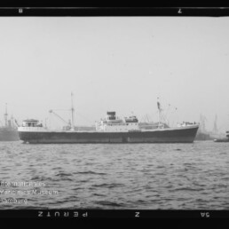 Frachtschiff im Hafen, von einem Schlepper begleitet, mit Hafenkränen im Hintergrund und Museumslogo unten links.