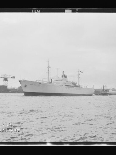 Großes weißes Schiff mit Masten und Aufbauten liegt im Hafen, im Hintergrund Werftkräne und ruhiges Wasser.