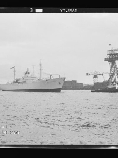 Weißes Schiff, begleitet von Schleppern, fährt an einem Werftgelände mit hohen Kranen und der Aufschrift 