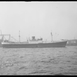 Seitliche Aufnahme eines Frachtschiffs im Hafen, Kräne und Werftgebäude im Hintergrund, Museumswasserzeichen links unten