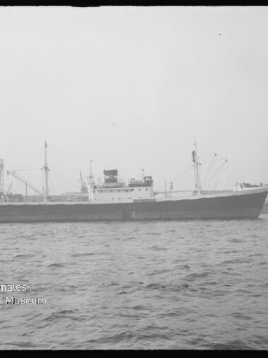 Seitliche Aufnahme eines Frachtschiffs im Hafen, Kräne und Werftgebäude im Hintergrund, Museumswasserzeichen links unten