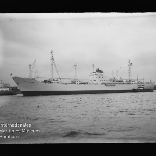 Weißes Passagiersschiff mit geschmückten Masten am Kai, Schlepper am Heck und Museumslogo links unten