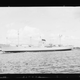 Weißes Frachtschiff mit mehreren Masten fährt auf ruhiger See, kleiner Schlepper am Heck.