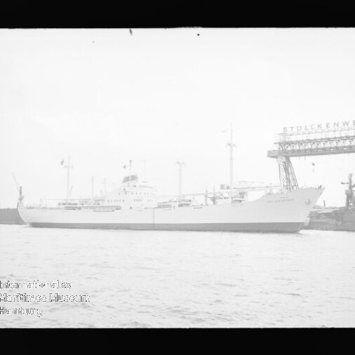 Langes weißes Passagierschiff im Hafen, neben einer Werft mit Kran und Pier sichtbar.