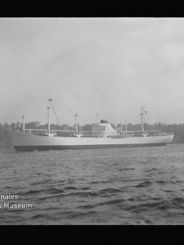 Frachtschiff seitlich auf ruhiger See, Masten und Ladegeschirr, Ufer mit Bäumen und Gebäuden; Museumslogo links unten.