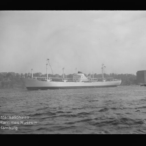 Frachtschiff seitlich auf ruhiger See, Masten und Ladegeschirr, Ufer mit Bäumen und Gebäuden; Museumslogo links unten.