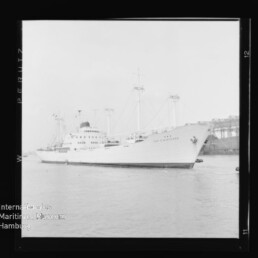 Weißes Frachtschiff mit mehreren Masten und Aufbauten liegt im Hafen vor einem Pier mit Kränen.