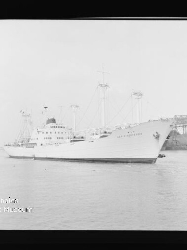 Weißes Frachtschiff mit mehreren Masten und Aufbauten liegt im Hafen vor einem Pier mit Kränen.