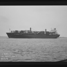Schwarzweißaufnahme eines beladenen Containerschiffs auf ruhiger See mit gestapelten Containern, seitliche Ansicht.