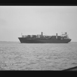 Containerschiff mit aufgestapelten Containern auf ruhiger See, schwarzweißes Foto mit Museumswasserzeichen unten links.