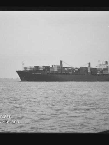 Containerschiff mit aufgestapelten Containern auf ruhiger See, schwarzweißes Foto mit Museumswasserzeichen unten links.