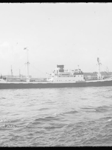 Frachtschiff mit mittigem Schornstein und zwei Masten vor Hafenkränen auf ruhiger See; Museumslogo links unten.