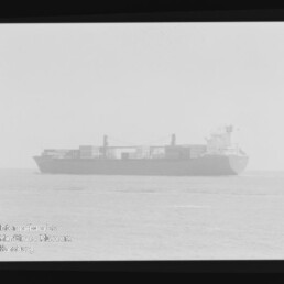 Frachtschiff mit gestapelten Containern fährt im Nebel auf ruhiger See; Filmrand und Museumswasserzeichen links unten.