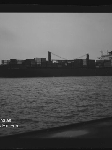 Schwarzweiß-Foto eines beladenen Containerschiffs mit zwei Ladekränen auf ruhiger See, Museumswasserzeichen unten links.