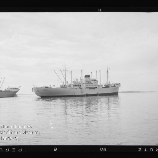 Großes Frachtschiff mittig auf ruhiger See, ein zweites Schiff links im Hintergrund, Schwarzweiß-Aufnahme.