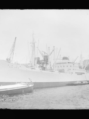 Großes weißes Passagier‑ und Frachtschiff am Kai eines Industriehafens, Krananlagen im Hintergrund und ein Schlepper im Wasser.