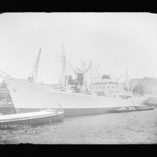 Großes weißes Passagier‑ und Frachtschiff am Kai eines Industriehafens, Krananlagen im Hintergrund und ein Schlepper im Wasser.