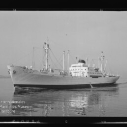 Großes weißes Frachtschiff mit Aufbauten, Masten und Raucherabzug auf ruhiger See, Spiegelung im Wasser und Museumslogo unten links.