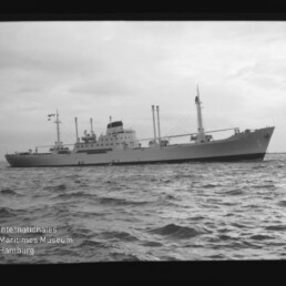 Großes Frachtschiff mit Masten und mittiger Aufbauten auf ruhiger See, links unten Wasserzeichen 