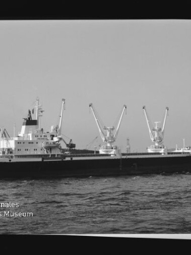 Frachtschiff mit drei Laderkränen auf dem Wasser vor Hafenanlagen; Wasserzeichen: Internationales Maritimes Museum Hamburg.