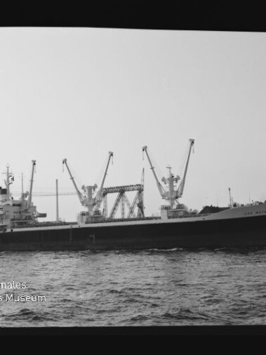 Frachtschiff mit drei hohen Ladekränen auf See vor einem Hafen, schwarz-weißaufnahme mit Museumslogo unten links.